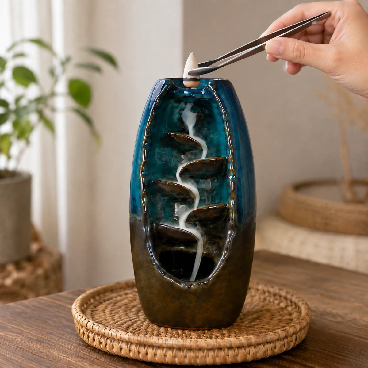 Blue ceramic vase with water flowing through stones on a wooden surface, with a hand using tongs placing incense cone.