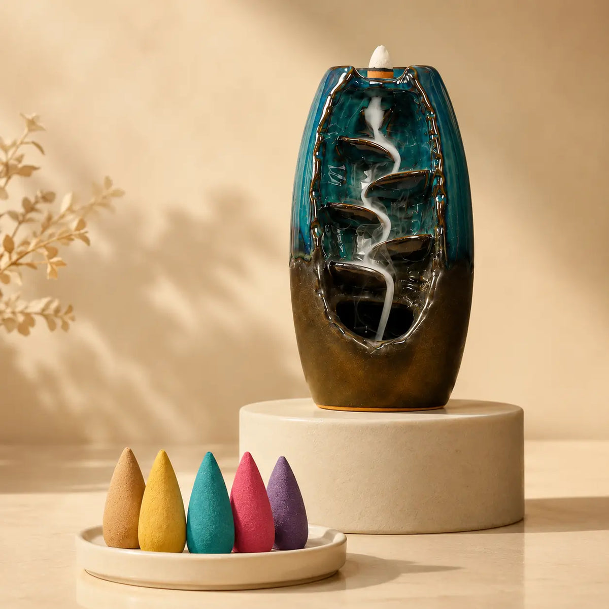 A ceramic backlit incense burner with a flowing white incense trail, displayed on a round beige pedestal. In the foreground, a small white dish holds five colorful incense cones in beige, yellow, teal, pink, and purple.