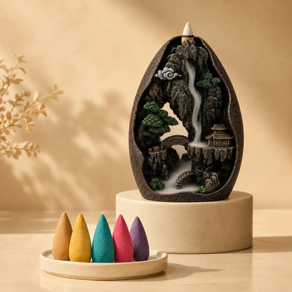 A decorative incense burner sculpture shaped like an egg, featuring a miniature mountain landscape with a waterfall, trees, bridge, and pagoda, displayed on a round pedestal. Five colorful incense cones in tan, yellow, teal, pink, and purple sit in a small dish in the foreground against a warm beige background.
